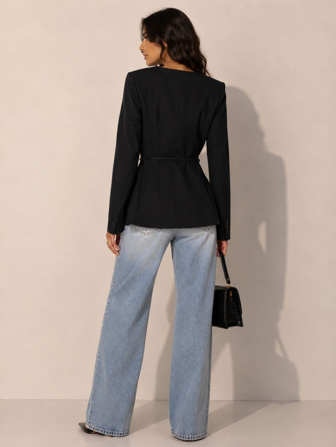 A woman shown from the back wearing a black belted long-sleeve blazer and wide-leg light blue jeans, holding a small black handbag against a plain beige studio background.