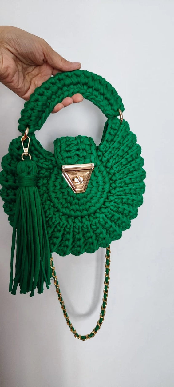 Green crocheted handbag with tassel and chain detail held by a hand against a white background