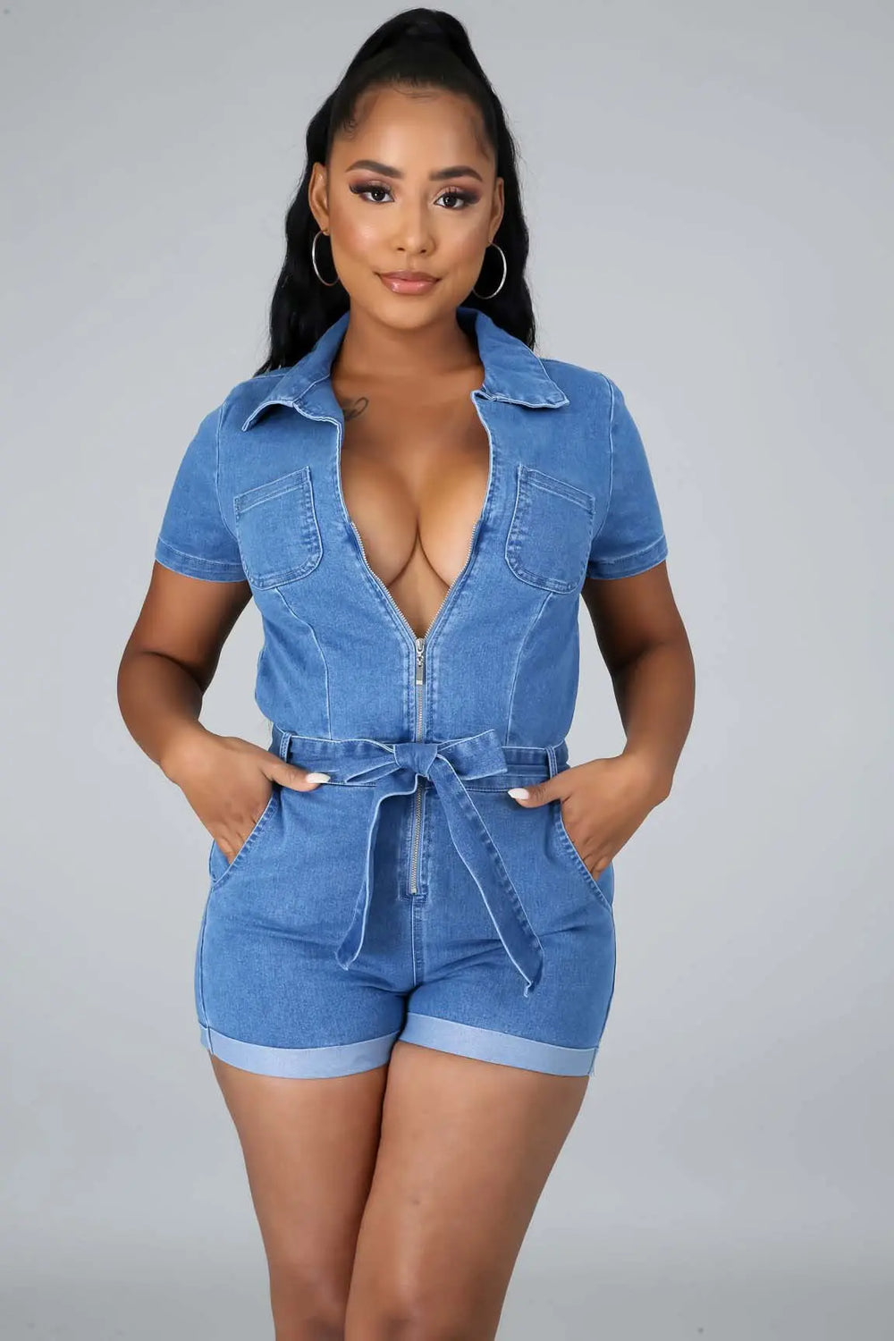 Front view of medium wash denim shorts romper with zipper front, collared neckline, tie waist belt and chest pockets on model