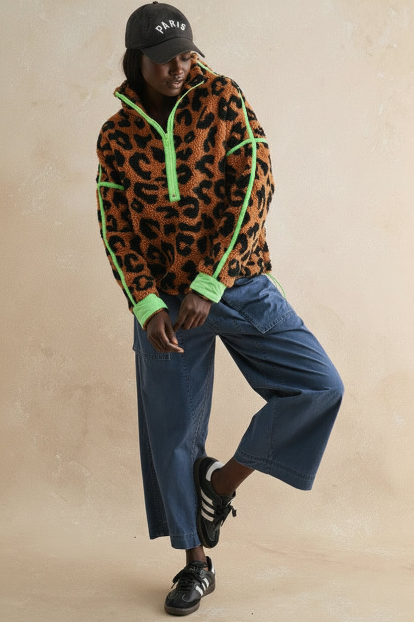A model wearing a brown and black leopard print fleece pullover with bright green trim, paired with wide-leg blue jeans, black sneakers, and a black baseball cap, posing against a textured beige wall.