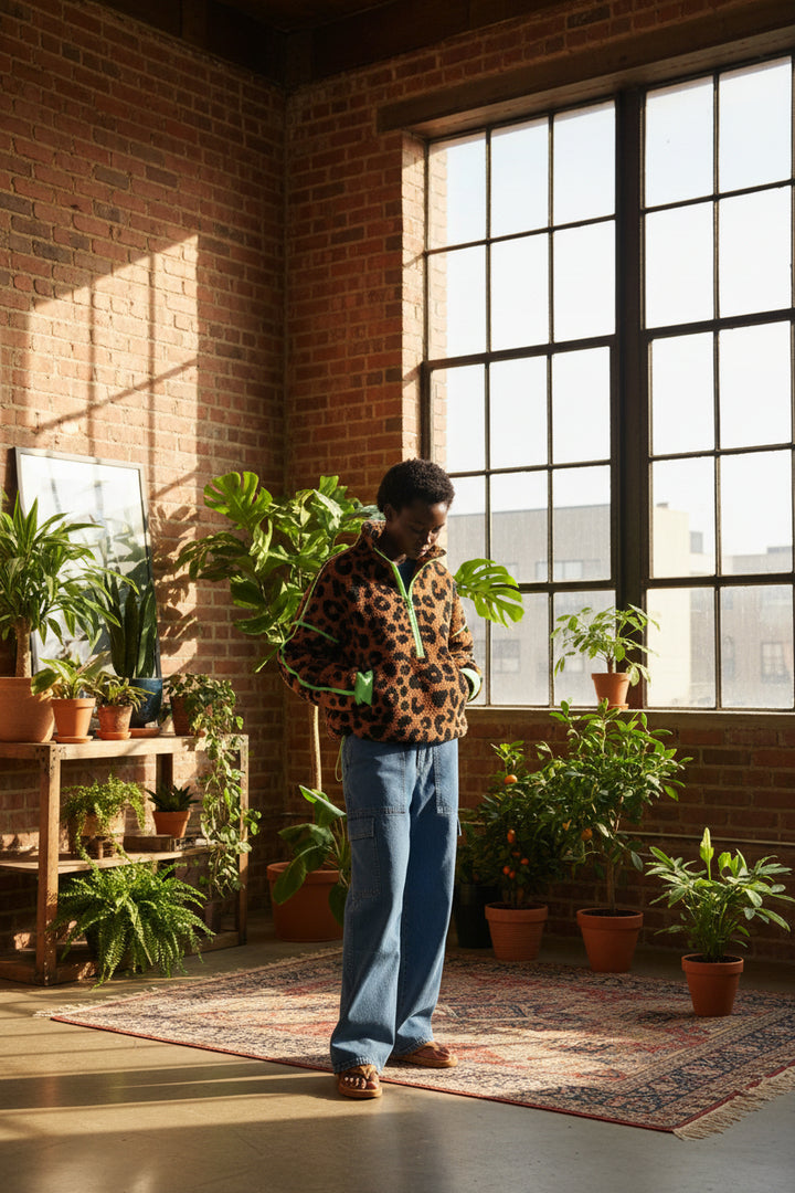 A model wearing a brown and black leopard print fleece jacket with bright neon green piping and cuffs, paired with blue jeans, set against a textured beige background.