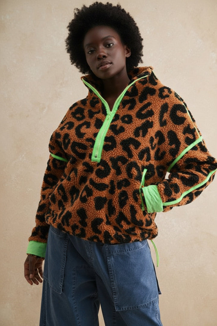 A model wearing a brown and black leopard print fleece pullover with bright neon green trim and a green zipper, paired with blue jeans, set against a textured beige background.