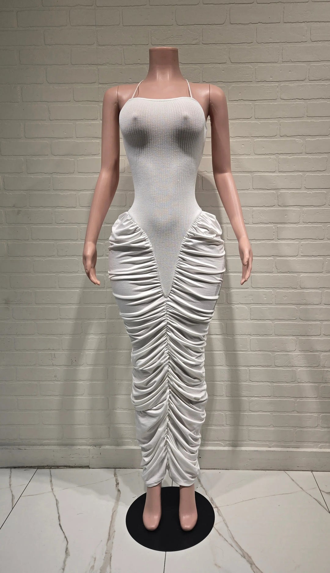 A sleeveless white bodycon dress displayed on a female mannequin against a white brick wall. The dress features a fitted bodice and dramatic vertical ruching and draping down the skirt, ending in a gathered ankle-length hem.